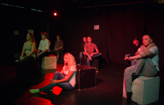Zdjęcie przedstawia scenę ze spektaklu. Grupa młodych aktorów siedzi na czarnych kubikach, oświetlenie jest czerwone.