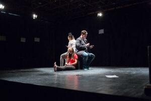 Zdjęcie przedstawia scenę ze spektaklu. Na scenie siedzi trójka młodych aktorów, dwójka z nich siedzi na czarnym podeście a trzecia siedzi z wyciągniętymi nogami, opierając się o ten podest.