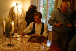 Zdjęcie przedstawia młodego aktora siedzącego przy stole, na którym leży ozdobna serweta, biała filiżanka, drewniane pudełko i kandelabr na trzy świece. Za mężczyzną jest okno z widokiem na ogród. Obok niego stoi kobieta i mężczyna, widzowie spektaklu.