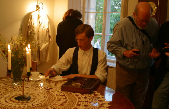 Zdjęcie przedstawia młodego aktora siedzącego przy stole, na którym leży ozdobna serweta, biała filiżanka, drewniane pudełko i kandelabr na trzy świece. Za mężczyzną jest okno z widokiem na ogród. Obok niego stoi kobieta i mężczyna, widzowie spektaklu.
