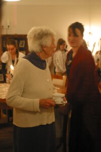 Zdjęcie przedstawia dwie kobiety patrzące na siebie. Kobieta po lewej stronie ma krótkie siwe włosy i trzyma filiżankę w ręku, kobieta po prawej ma ciemne włosy upięte w kok, ubrana jest w strój z epoki stylizowany na lata 40 XX wieku. Za nimi w głębi pomieszczenia widać inne osoby.