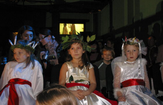 Zdjęcie przedstawia trzy dziewczynki ubrane na biało w sukienki przepasane czerwoną szarfą. Na głowach mają wianki z zielonych liści. Dziewczynka po lewej stronie ma na głowie również wianek ze świec.