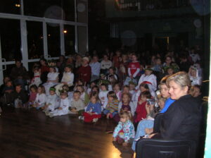 Zdjęcie przedstawia widowsnię siedzącą na podłodze i na krzesłach. W dużej części widownia składa się z dzieci.