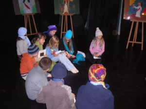 Zdjęcie przedstawia grupę dzieci siedzących na podłodze w półogręgu na scenie. Ubrani są w kolorowe bluzy i swetry, na głowach mają zimowe czapki.