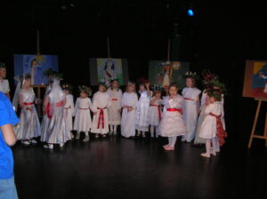 Zdjęcie przedstawia grupę dziewczynek na scenie ubranych w białe sukienki przepasane czerwonymi wstążkami. Dziewczynki stoją w półokręgu i mjaą na głowach wianki z zielonych liści.
