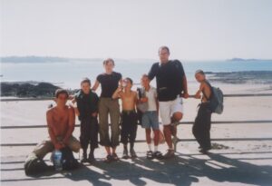 Zdjęcie grupowe przedstawia 7 osób - dorosłych i dzieci, opartych o barierki. W tle widać morze i wyspy oraz szeroką plażę. Dzień jest ciepły i słoneczny.