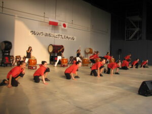 Zdjęcie grupowe zrobione na dużej hali sportowej. Na ścianie widzą dwie flagi: jedna biało-czerwona i druga biała z czerwonym okręgiem na środku. Na sali jest grupa młodych osób robiących pokaz. Część z nich ubrana w pomarańczowo-czerwone koszulki i czarne spodnie kuca przy podłodze w tej samej pozie. W głębi pod ścianą jest rząd osób grających na dużych japońskich bębnach. Ubrani są w czarne koszulki i białe spodnie.