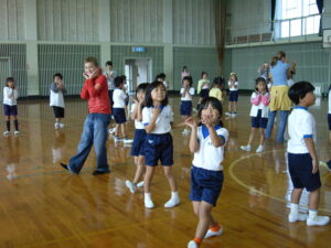 Zdjęcie przedstawia dużą grupę dzieci na sali gminastycznej. Dzieci z azjatyckimi rysami twarzy ubrane są w biało-granatowe mundurki szkolne i robią miny do zdjęcia. Wśród nich jest paru nastolatków o europejskich rysach twarzy w kolorowych bluzach i jeansach.