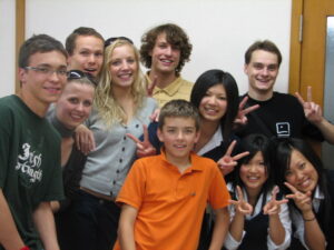 Zdjęcie grupowe zrobione w pomieszczeniu, białe tło. Na zdjęciu jest 9 osób, dziewcząt i chłopców, Europejczyków i Azjatów. Wszyscy uśmiechają się do zdjęcia.