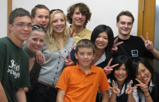 Zdjęcie grupowe zrobione w pomieszczeniu, białe tło. Na zdjęciu jest 9 osób, dziewcząt i chłopców, Europejczyków i Azjatów. Wszyscy uśmiechają się do zdjęcia.