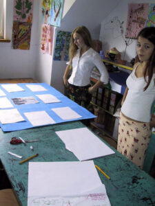 Zdjęcie przedstawia dwie nastolatki stojące nad stołem w pracowni plastycznej. Na ścianach wiszą prace a przed nimi na stole leży niebieska plansza z białymi kartkami do niej przyklejonymi.