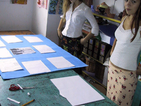 Zdjęcie przedstawia dwie nastolatki stojące nad stołem w pracowni plastycznej. Na ścianach wiszą prace a przed nimi na stole leży niebieska plansza z białymi kartkami do niej przyklejonymi.
