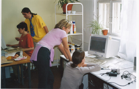 Zdjęcie przedstawia dwie kobiety przy monitorze komputerowym, jedna z nich kuca przy biurku, druga stoi opierając się o biurko. W tle stoi mężczyzna z długimi włosami w żółtej koszulce i krawacie stoi za chłopcem pracującym przy drugim komputerze..