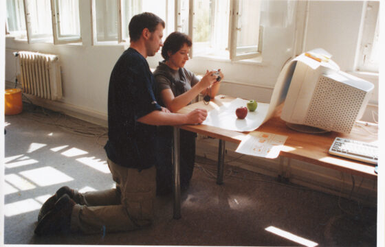 Dwie osoby - kobiera i mężczyzna, klęczą przed biurkiem z dużym monitorem komputerowym. Kobieta trzyma w ręku aparat fotograficzny.