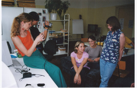 Zdjęcie przedstawia 5 kobiet w pomieszczeniu. Dwie po lewej stronie siedzą na stole i trzymają w rękach kamery, dwie kucają na środku zdjęcia, a po prawej stronie jedna stoi patrząc na grupę.