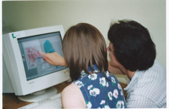 Dwie kobiety siedzą tyłem patrząc na ekran dużego monitora komputerowego. Jedna z nich pokazuje palcem na ekran.