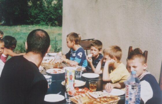 Zdjęcie przedstawia grupę dzieci z dorosłym mężczyzną jedzących obiad przy stole w ogrodzie.