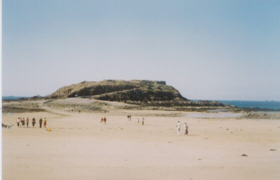 Zdjęcie przedstawia szeroką i dużą plażę, w oddali widać sylwetki ludzi. Plaża kończy się wzniesieniem. Na horyzoncie widać błękitne morze.