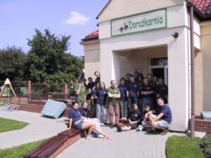 Zdjęcie grupowe przed wejściem do budynku Dorożkarni. Grupa osób siedzi i stoi w centarlnej części zdjęcia. Nad nimi na fasadzie budynku wisi kolorowy napis Dorożkarnia.