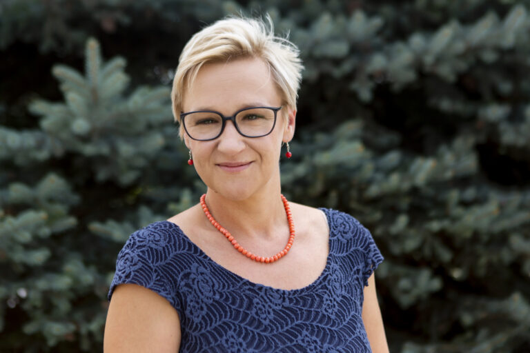 Zdjęcie przedstawia młodą kobietę. Jest krótko ostrzyżona, ma korale na szyi i okulary na nosie.