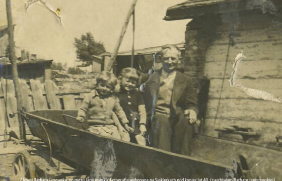 To jest bardzo stare zdjęcie, z pewnością pochodzi z wczesnych lat po II wojnie światowej. Przedstawia mężczyznę zdójkę dzieci siedziących na wozie. Za nimi widzimy fragment drewnianego domu.