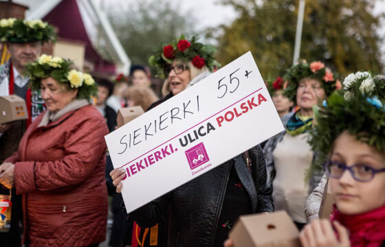 Grupa ludzi stojących na dworzu. Na głowach mają wianki. Kobieta w środku trzyma planszę z napisem Siekierki 55+ Siekierki Ulica Polska.