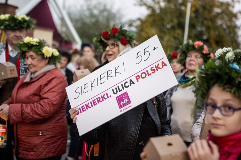 Grupa ludzi stojących na dworzu. Na głowach mają wianki. Kobieta w środku trzyma planszę z napisem Siekierki 55+ Siekierki Ulica Polska.