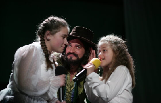 Na zdjęciu dwie dziewczynki, między nimi mężczyzna w kapeluszu. Dziewczynka z prawej trzyma w dłoni mikrofon.