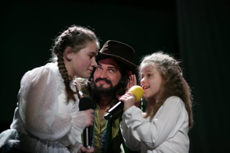Na zdjęciu dwie dziewczynki, między nimi mężczyzna w kapeluszu. Dziewczynka z prawej trzyma w dłoni mikrofon.
