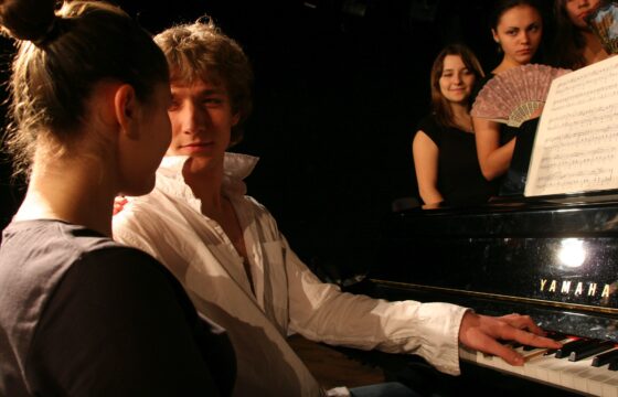 |Zdjęcie: scena, przy fortepianie siedzą chłopak i dziewczyna, za fortepianem grupa dziewczyn z wachlarzami w dłoniach