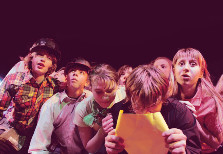 Grupa młodzieży na scenie, dziewczęta i chłopcy, stoją blisko siebie, ściśnięci. Chłopak nw pierwszym rzędzie patrzy na kartkę, którą trzyma w dłoniach blisko twarzy.