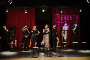 Osiem młodych osób na scenie w kostiumach z lat dwudziestych ubiegłego wieku. Za nimi napis Seventh Heaven