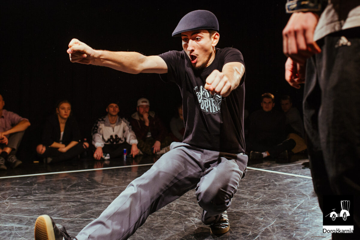 Młody chłopak na tańczy, najprawdopodobniej breakdance. Wokół niego tłum młodych ludzi.