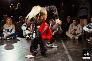 Młoda dziewczyna tańczy prawdopodobnie breakdance, długie włosy ma związane w koński ogon, wokół niej na podłodze siedzą młodzi ludzie