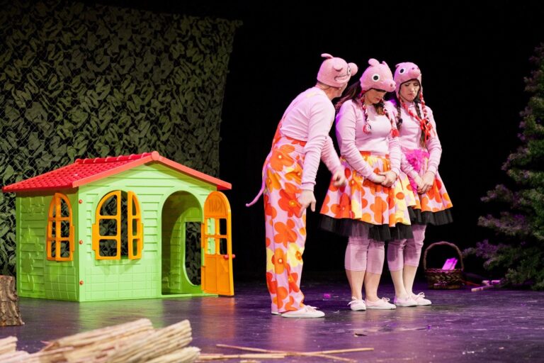 Trzy postacie na scenie w kostiumach świnek, za nimi mały domek