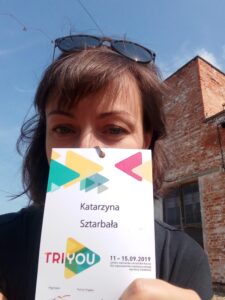 Zdjęcie przedstawia kobietę z ciemnymi włosami, grzywką i okularami na czubku głowy. Twarz do połowy zasłonięta jest identyfikatorem konferencji TRIYOU. W tle widać fragment ceglanego budynku