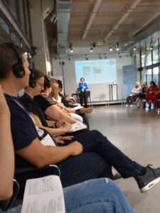 Zdjęcie przedstawia salę konferencyjną. Po lewej stronie siedzą osoby w słuchawkach, w tle widać projekcję prezentacji i osobę prowadzącą.