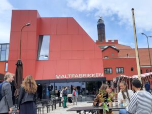 Zdjęcie przedstawia front budynku fakbryki słodu. Fasada jest pomarańczowa z napisem Maltfabrikken. Na placu przed budynkiem widać rozmawiające osoby zgromadzone wokół stolików.