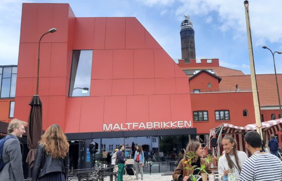 Zdjęcie przedstawia front budynku fakbryki słodu. Fasada jest pomarańczowa z napisem Maltfabrikken. Na placu przed budynkiem widać rozmawiające osoby zgromadzone wokół stolików.