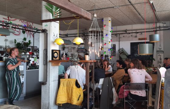 Zdjęcie przedstawia wnętrze kawiarni z grafiti na ścianach. Przy stolikach siedzą osoby. Po lewej stronie stoi kobieta i mówi do grupy.