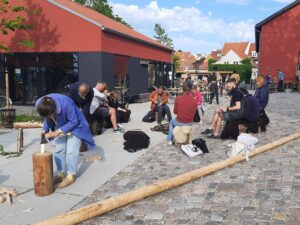 Zdjęcie przedstawia grupę osób pracujących przy drewnie na świeżym powietrzu, między dwoma budynkami. Każdy indywidualnie rzeźbi kawałki drewna. Po prawej stronie na dole leży długi drewniany pal.