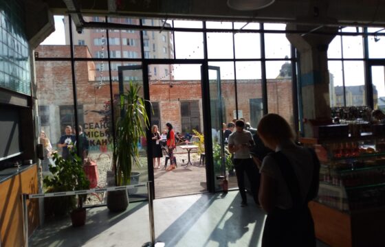 Zdjęcie przedstawia wnętrze budynku i kawiarnię. Ściana w tle jest ze szkła, w niej otwarte drzwi prowadzące na taras. Po prawej stronie oraz na tarasie widać zgromadzone osoby.