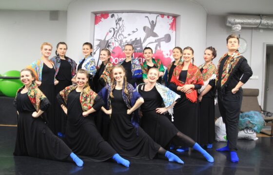 Zdjęcie grupowe zespołu tanecznego. Wszyscy ubrani są w czarne sukienki i spodnie, na stopach wszyscy mają intensywnie niebieskie skarpetki. Na ramionach wszyscy mają kolorowe, ludowe chusty.