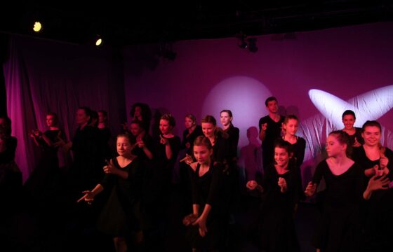 Zdjęcie przedstawia fragment spektaklu. Duża grupa dziewcząt w czarnych sukienkach tańczy na scenie. Światła są czerwono-wiśniowe, nie widać dokładnie wszystkich sylwetek i twarzy.