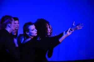 Zdjęcie przedstawia fragment spektaklu. Dwie kobiety wyciągają ręce w tym samym kierunku. Za nimi stoją dwie tancerki. Tło jest niebieskie, kobiety ubrane są na czarno.