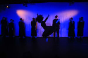 Zdjęcie przedstawia fragment spektaklu. Siedem osób stoi w linii na końcu sceny, na pierwszym planie widać skaczącą kobietę rozpościerającą ręce. Światło jest niebieskie, słabe.
