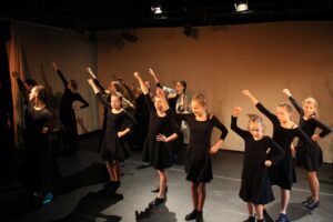 Fragment spektaklu. Grupa dziewcząt w czarnych sukienkach i związanych włosach stoi na scenie z uniesioną prawą ręką.