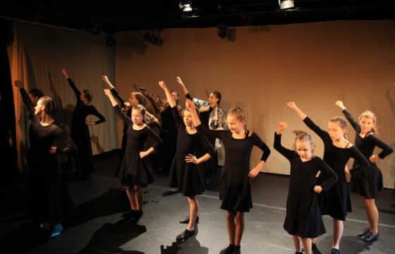Fragment spektaklu. Grupa dziewcząt w czarnych sukienkach i związanych włosach stoi na scenie z uniesioną prawą ręką.
