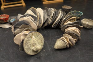 Zdjęcie przedstawia pracę ceramiczną w kształcie łańcucha składającego się z płaskich, okgrągłych elementów w kolorach ziemi.