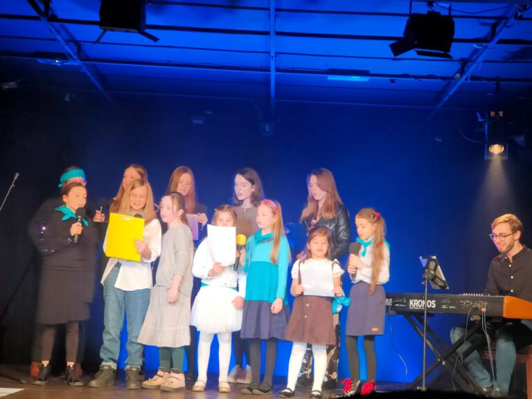 Grupa dzieci i młodzieży na scenie - 12 dziewcząt, z prawej strony chłopak gra na keyboardzie